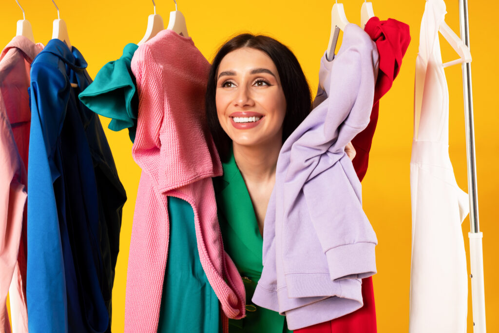 Smiling beautiful european woman posing between colorful new clothes hanging on garment rack, yellow
