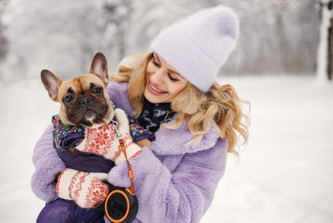 Woman in knitted hat playing with her dog french bulldog on a snow