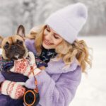 Woman in knitted hat playing with her dog french bulldog on a snow